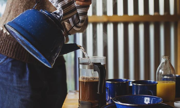 A close up of a person pouring hot water into a cafetière full of coffee. Also a bottle of organise juice and various cutlery.